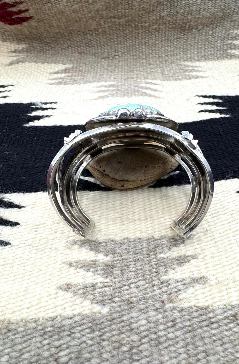 May include: Silver cuff bracelet with a large, oval turquoise stone set in a decorative bezel. The bracelet has a layered design and rests on a neutral-toned patterned textile.