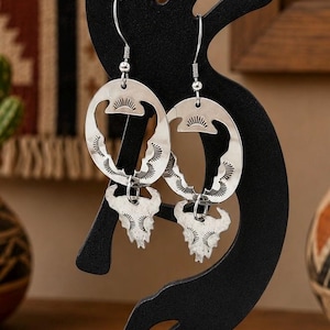 May include: A pair of silver-toned dangle earrings featuring a circular hoop design with stamped sunburst patterns and a dangling bull skull charm. The earrings are suspended from silver-toned metal hooks and display a polished, metallic finish.