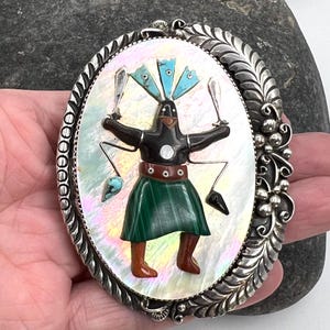 May include: Oval silver-tone pendant with a central design featuring a figure with a black top, green skirt, and brown boots. The figure holds two silver objects and is surrounded by a mother-of-pearl background. The pendant has a decorative silver border.