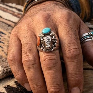 May include: A silver ring with a turquoise stone and a buffalo head design. The ring also features a coral-colored stone. Another ring with a turquoise stone is visible on the adjacent finger. The rings are on a hand.