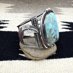 May include: A silver cuff bracelet with a large, oval turquoise stone. The bracelet features a decorative feather design and geometric patterns. The turquoise stone is a light blue color with brown matrix. The bracelet is displayed on a small, gray stone.