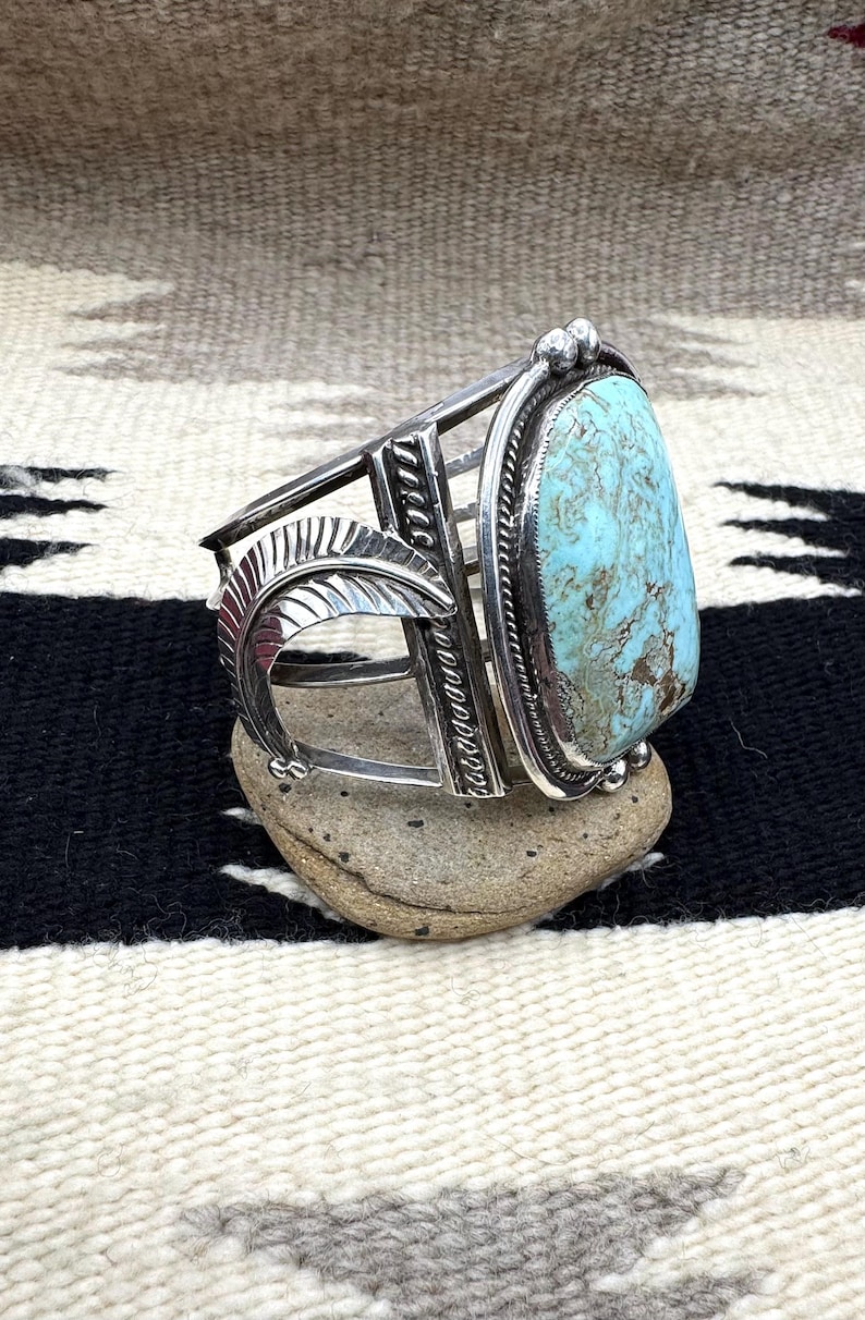 May include: A silver cuff bracelet with a large, oval turquoise stone. The bracelet features a decorative feather design and geometric patterns. The turquoise stone is a light blue color with brown matrix. The bracelet is displayed on a small, gray stone.