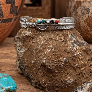 May include: A silver cuff bracelet shaped like a feather with a wrapped wire detail at one end. The bracelet features five small, round, bezel-set cabochon stones in turquoise, green, white, black, and red, arranged in a row along the center.