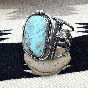 May include: A silver cuff bracelet with a large, rectangular turquoise stone. The stone is set in a detailed silver frame with decorative elements, including small silver beads and leaf designs. The bracelet is displayed on a small stone.