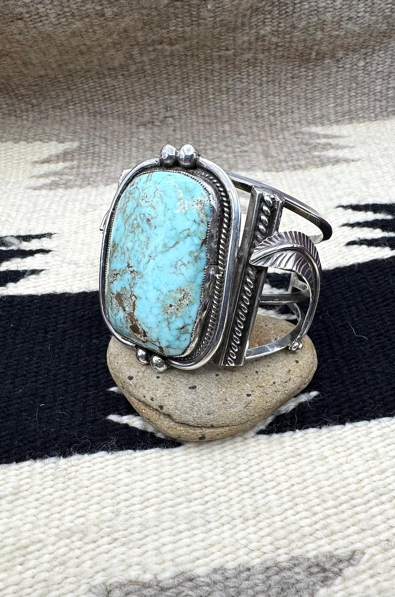 May include: A silver cuff bracelet with a large, rectangular turquoise stone. The stone is set in a detailed silver frame with decorative elements, including small silver beads and leaf designs. The bracelet is displayed on a small stone.