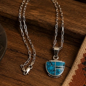 May include: A silver chain necklace featuring a semi-circular pendant inlaid with blue turquoise stone. The pendant is divided into sections by thin silver lines, with one side displaying a vibrant turquoise pattern and the other side featuring black horizontal stripes.