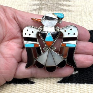 May include: A silver-toned pendant in the shape of a bird, featuring inlaid stones in black, white, turquoise, and orange. The bird has a detailed design with a geometric pattern and is suspended from a delicate chain.