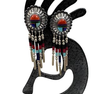 May include: Silver-toned dangle earrings with a sunburst design, featuring a central oval of red, blue, and turquoise stones. The earrings have beaded strands in red, black, and blue, with silver-toned fringe.