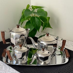 May include: A vintage silver tea set with wooden handles and lids, including a teapot, creamer, sugar bowl, and a small teapot, all on a rectangular silver tray. The set is displayed on a black patterned runner.