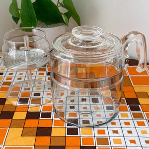 May include: A clear glass coffee pot with a lid and handle, alongside a clear glass carafe. The coffee pot is cylindrical with horizontal ridges. The background features a plant in a white pot and a patterned orange, brown, and white tablecloth.