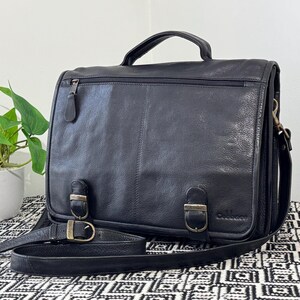 May include: Black leather briefcase with a top handle, shoulder strap, and metal buckle fastenings. The bag has a zipped pocket on the front and a zipped compartment on the back. The bag is on a patterned surface.