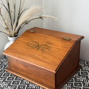 May include: A wooden slant-top desk box with a hinged lid featuring a dark green painted floral motif. The box has a rectangular base, a smooth medium-brown wood grain finish, and two visible brass-coloured metal hinges on the top edge.