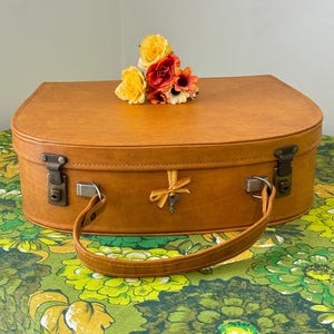 May include: A vintage tan suitcase with a curved top, metal latches, and a leather handle. A small bouquet of yellow, orange, and red flowers rests on top. The suitcase has a key tied with a gold ribbon.