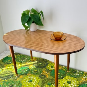 May include: An oval wooden coffee table with four tapered legs featuring gold-tone metal feet. On the table sits a small amber-coloured glass teacup and saucer, and a green leafy plant in a white ceramic pot.