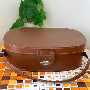 May include: A brown oval vintage suitcase with a handle and a metal clasp. The suitcase is made of a smooth material and has a stitched design around the edges. The handle is attached to the side. The suitcase is on a patterned surface.