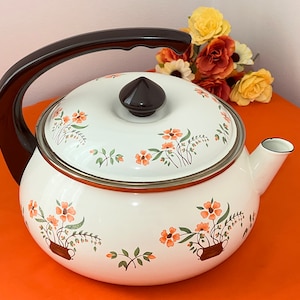 May include: White enamel teapot with a brown handle, lid, and floral design. The teapot features orange flowers and green leaves in brown pots. The spout and rim are trimmed in silver. A vintage kitchenware item.