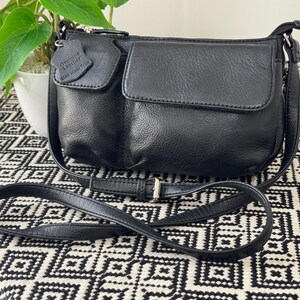 May include: Black leather crossbody bag with a flap pocket and a small tag. The bag has a long, adjustable strap and a zip closure. The bag is on a black and white patterned surface.