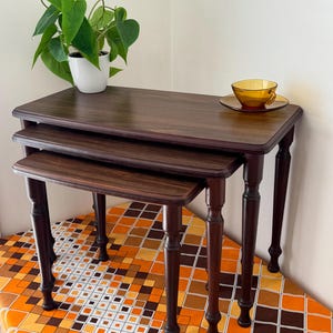 May include: A set of three nesting tables with dark brown wood grain surfaces and turned legs. The tables are stacked in a descending arrangement. A white pot with a green plant and an amber glass teacup with a saucer sit on the largest table.