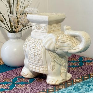 May include: A cream-coloured elephant garden stool with detailed carvings. The elephant has a raised trunk and decorative patterns. The stool sits on patterned fabric, with a white vase and dried plants in the background. The stool is a decorative item.