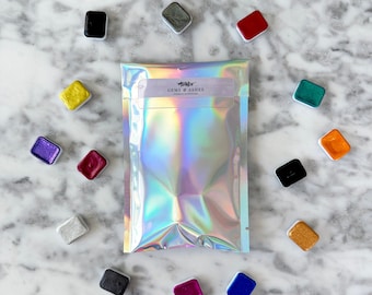 May include: A collection of watercolor paints in various colors, including black, yellow, purple, and red, arranged around a holographic pouch. The pouch has the text "GEMS & ASHES" printed on it. The paints are in small, square pans.