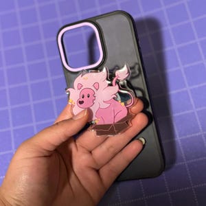 Könnte beinhalten: Ein rosa Löwen-Cartoon-Charakter in einer Box, befestigt an einer schwarzen Handyhülle mit einem lila Rand. Der Löwe hat eine flauschige Mähne und einen Schwanz mit kleinen gelben Sternakzenten. Der Hintergrund ist ein lila Gitter.