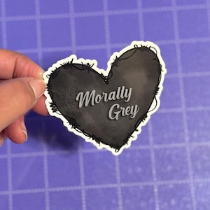 May include: A heart-shaped sticker with the words "Morally Grey" in a cursive font. The heart is filled with a dark gray watercolor effect and outlined with a barbed wire design. The sticker is held by a hand.