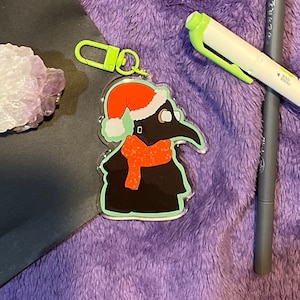 May include: A green and red Christmas-themed keychain featuring a cartoon plague doctor wearing a Santa hat and a red scarf.