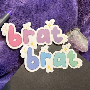May include: Two stickers with the word "brat" written in a bubbly font. The top sticker is pink, purple, and blue. The bottom sticker is blue, green, and light blue. Both stickers have gold glitter accents.