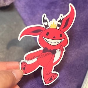May include: Red cartoon devil with a gold crown, black bow tie, and black hearts on its feet. The devil is smiling with sharp teeth and has a tail.