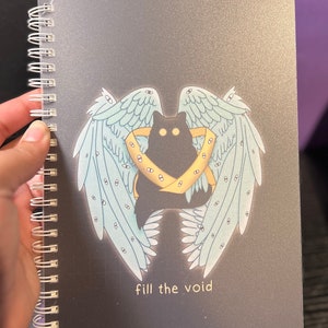 K&ouml;nnte beinhalten: Eine schwarze Katze mit gelben Augen ist von blauen Fl&uuml;geln mit Augen darauf umgeben. Die Fl&uuml;gel befinden sich auf einem schwarzen Hintergrund mit dem Text "fill the void" in Gelb.