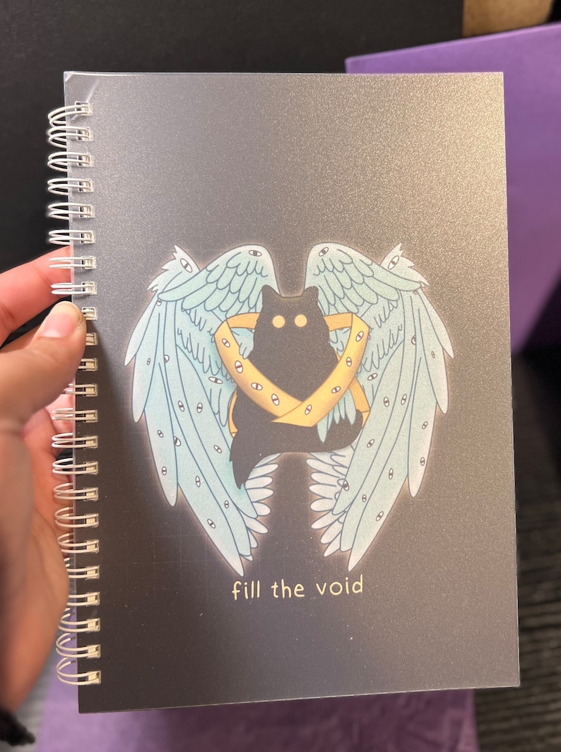 K&ouml;nnte beinhalten: Eine schwarze Katze mit gelben Augen ist von blauen Fl&uuml;geln mit Augen darauf umgeben. Die Fl&uuml;gel befinden sich auf einem schwarzen Hintergrund mit dem Text "fill the void" in Gelb.