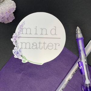 Könnte beinhalten: Runder weißer Aufkleber mit dem Text "mind matter" in grauer Schrift mit einer Linie, die die Wörter trennt. Der Aufkleber hat ein florales Design mit lila Blumen und grünen Blättern um den Text herum.