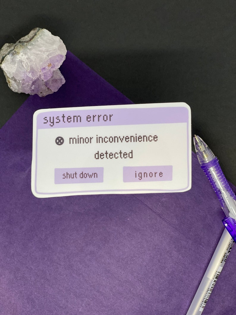 Puede incluir: Una pegatina morada y blanca con el texto "system error" y una se&ntilde;al de advertencia. El texto "minor inconvenience detected" est&aacute; debajo de la se&ntilde;al de advertencia. Hay dos botones debajo del texto: "shut down" y "ignore".