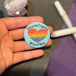 May include: A heart-shaped button with a rainbow design and the text "we aren't going anywhere".