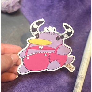 May include: A sticker depicting a cartoon creature with purple skin, pink dungarees, and a yellow beak. The creature has black and white striped horns and a tag with the word "THING?" on it.