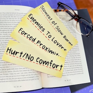 May include: Four yellow index cards with black text, stacked on an open book. The cards read "Slowest of Slow Burns," "Enemies To Lovers," "Forced Proximity," and "Hurt/No Comfort." Eyeglasses rest on the book. The theme is romance.