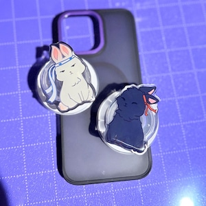 Könnte beinhalten: Zwei Handyhalterungen mit Cartoon-Hasen-Designs. Einer ist weiß mit einem blauen Stirnband, der andere ist schwarz mit einer roten Schleife. Beide befinden sich auf einer schwarzen Handyhülle mit einem lila Kameraring.