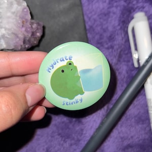 May include: A green button with a cartoon frog drinking water from a glass. The button says "hydrate stinky" in a handwritten font.