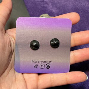 May include: A close-up of the back of two black enamel pins with a purple and gray gradient background. The background has the text "@CHAOTICPOSSUMDESIGNS" and social media icons for TikTok, Instagram, and email.