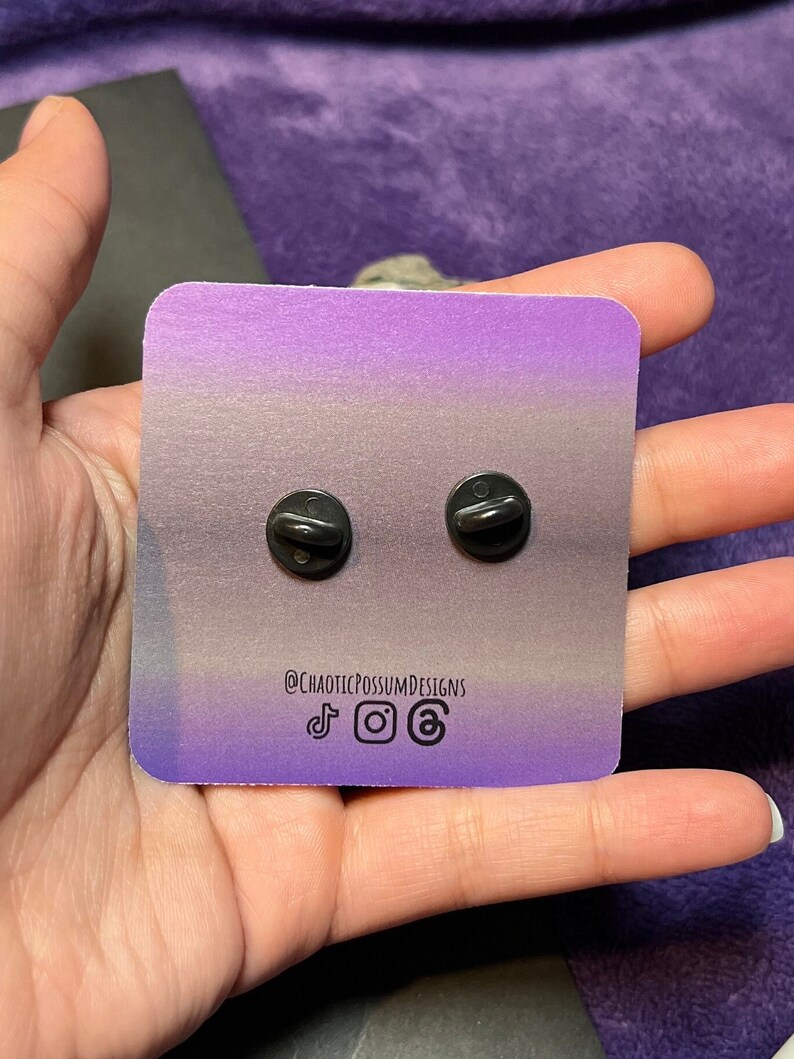May include: A close-up of the back of two black enamel pins with a purple and gray gradient background. The background has the text "@CHAOTICPOSSUMDESIGNS" and social media icons for TikTok, Instagram, and email.