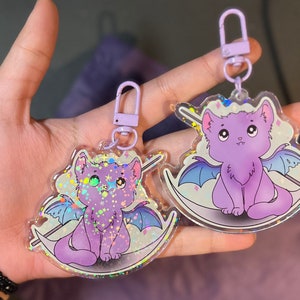 Pu&ograve; includere: Due portachiavi a forma di gatto color viola iridescente con dettagli glitterati. Un gatto &egrave; un gatto nero con le ali e l'altro &egrave; un gatto bianco con le ali. Entrambi i gatti sono seduti su una luna crescente.