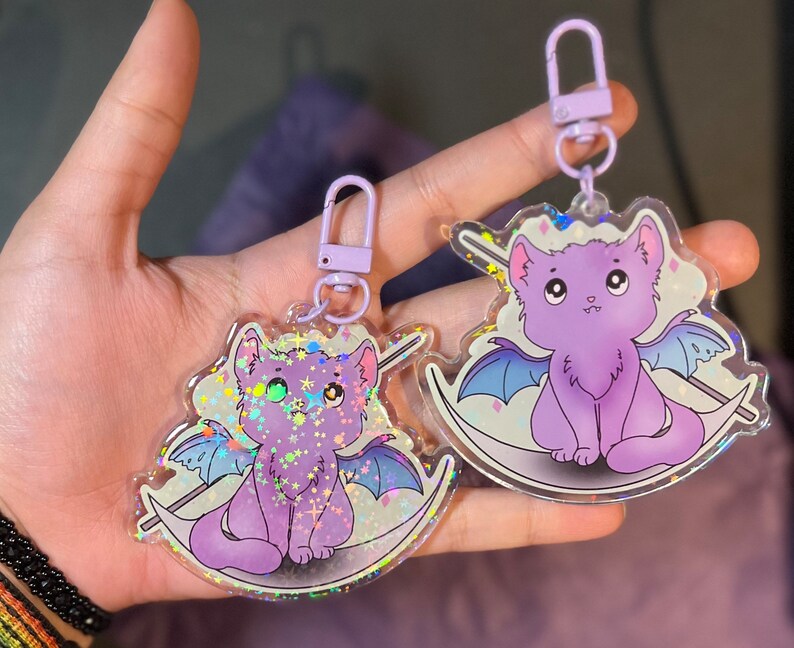 Pu&ograve; includere: Due portachiavi a forma di gatto color viola iridescente con dettagli glitterati. Un gatto &egrave; un gatto nero con le ali e l'altro &egrave; un gatto bianco con le ali. Entrambi i gatti sono seduti su una luna crescente.