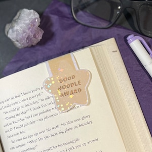 May include: A gold star-shaped bookmark with the text "GOOD NOODLE AWARD" in a glittery font. The bookmark is decorated with iridescent glitter and stars.