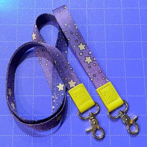 May include: Purple lanyard with a repeating pattern of white stars and dots. The lanyard has yellow leather accents and silver-colored metal clasps. The lanyard is designed to hold keys or an ID badge.