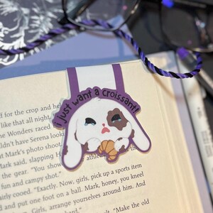 May include: Purple bookmark with a white bunny holding a croissant. The bunny has brown spots and is crying. The bookmark has the text "I just want a croissant!" on it.