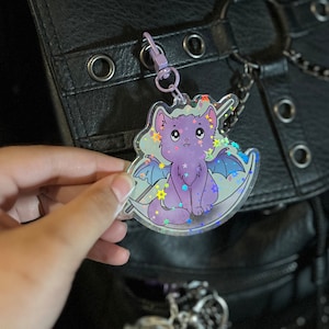 Pu&ograve; includere: Un ciondolo olografico con un gatto viola con le ali e una luna crescente. Il ciondolo &egrave; attaccato a una catena d'argento e ha una finitura scintillante.