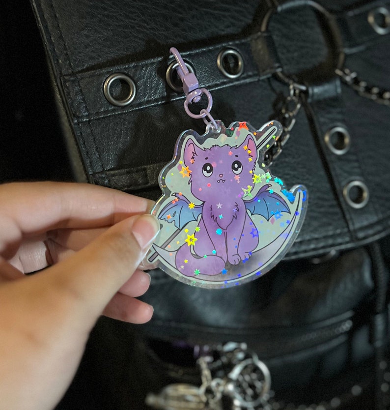 Pu&ograve; includere: Un ciondolo olografico con un gatto viola con le ali e una luna crescente. Il ciondolo &egrave; attaccato a una catena d'argento e ha una finitura scintillante.