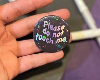 以下が含まれることがあります： ホログラフィックアクセントと「Please do not touch me」の文字が入った丸い黒いボタン。テキストは、色の組み合わせが楽しい遊び心のあるフォントです。ボタンは光沢のある反射面を持っています。