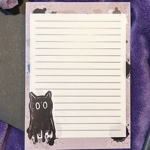 Puede incluir: Un bloc de notas blanco con líneas y una ilustración de un gato negro en la parte delantera. El gato tiene ojos grandes y gotea pintura negra. El fondo es una acuarela púrpura con salpicaduras de pintura negra.