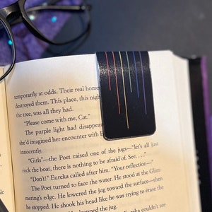 Puede incluir: Primer plano de un libro con un marcapáginas negro con rayas de colores. El texto en la página dice "Girls - the Poet raised one of the jugs - "let's all just rock the boat, there is nothing to be afraid of. See..."  "Don't!" Eureka called after him. "Your reflection -"